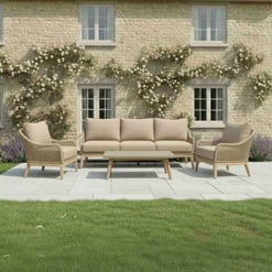 Stylish outdoor patio furniture set with beige cushions, surrounded by lush green lawn and flower beds in front of a charming brick house with blooming climbing roses.