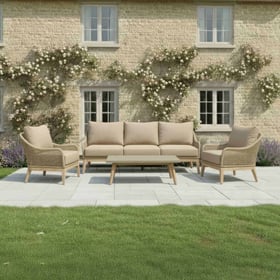 Stylish outdoor patio furniture set with beige cushions, surrounded by lush green lawn and flower beds in front of a charming brick house with blooming climbing roses.
