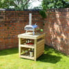 Portable stainless steel outdoor pizza oven with chimney on wooden cart in a cozy garden setting with brick wall and potted flowers.