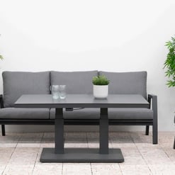 Lounge outdoor patio furniture set with grey cushioned chairs and sofa, surrounded by lush green plants and flowering shrubs for a stylish garden or backyard entertainment space.