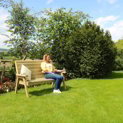 Relaxing woman sitting on a wooden garden bench in a lush green backyard with trees, grass, and scenic landscape.