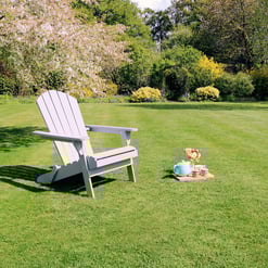 Adorable white Adirondack chair on lush green lawn with picnic setup, surrounded by blooming trees and vibrant garden, perfect for outdoor relaxation and garden decor.
