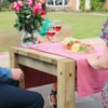 Vibrant outdoor patio with wooden dining table, pink checkered tablecloth, fresh cheese, charcuterie, cherry tomatoes, and two glasses of rosé wine, surrounded by lush green lawn and garden.