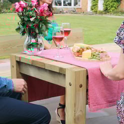 Vibrant outdoor patio with wooden dining table, pink checkered tablecloth, fresh cheese, charcuterie, cherry tomatoes, and two glasses of rosé wine, surrounded by lush green lawn and garden.