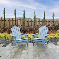 Bright outdoor garden seating with two blue Adirondack chairs on a paved patio, lush landscaping, and a wooden privacy fence, perfect for relaxing in a beautifully designed backyard garden.
