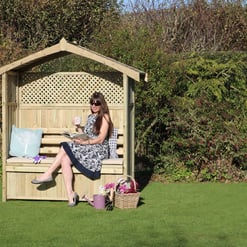 Rustic wooden garden bench with a shaded canopy, perfect for outdoor relaxation. Features decorative pillows and a gardener's basket with flowers, ideal for garden lounging and outdoor leisure.