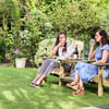 Relaxing outdoor garden dining scene with two women enjoying wine on wooden Adirondack chairs among lush greenery and blooming flowers. Perfect for outdoor furniture, garden decor, and relaxation.