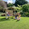 Kids playing in a lush, green backyard garden with a wooden summerhouse, surrounded by vibrant flowering bushes and trees, creating a perfect outdoor family space.