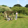 Relaxing outdoor garden scene with two women, one sitting on a wooden swing and the other standing beside it, surrounded by lush greenery and blooming plants.