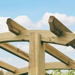 Sturdy wooden frame under construction for a garden arbor or pergola, featuring curved and angled beams, against a bright blue sky with scattered clouds.
