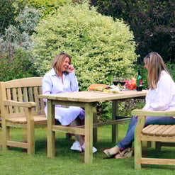 Relaxing outdoor garden dining scene with women enjoying food and drinks on wooden patio furniture amid lush greenery and colorful plants.