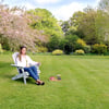 Bright young woman relaxing on adirondack chair in lush garden with blooming trees and vibrant green grass, perfect for outdoor living and gardening enthusiasts.