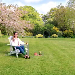 Bright young woman relaxing on adirondack chair in lush garden with blooming trees and vibrant green grass, perfect for outdoor living and gardening enthusiasts.