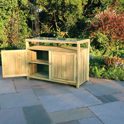Natural wood outdoor garden storage cabinet with glass tabletop on a stone patio amidst lush garden greenery.