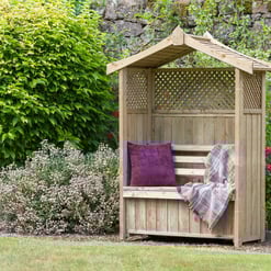 Cozy wooden garden gazebo with purple cushion and throw blanket surrounded by lush greenery and vibrant flowers for outdoor relaxation and garden decor.