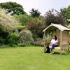 A woman sits on a wooden garden bench with a small shelter in a lush, colorful garden surrounded by blooming trees and shrubs. Perfect for outdoor relaxation and garden decoration.