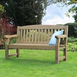 A wooden garden bench with a light blue cushion on lush green lawn surrounded by vibrant garden plants and trees.