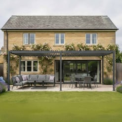 Modern backyard patio with outdoor seating, dining area, and pergola, featuring lush landscaping and blooming climbing roses against a classic stone house for outdoor living and garden enhancement.
