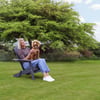 Relaxing outdoor garden scene with woman sitting on Adirondack chair, holding a dog under a large lush tree, surrounded by vibrant greenery and flowering plants.