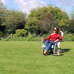 Serene woman relaxing outdoors in a lush garden on a sunny day, sitting in a comfortable Adirondack chair with a glass of white wine, surrounded by vibrant greenery and blooming trees.
