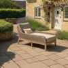 Luxurious outdoor chaise lounge with beige cushion on stone patio surrounded by lush garden and classic brick house, perfect for relaxing in a charming home and garden setting.