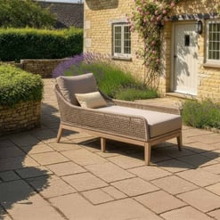 Luxurious outdoor chaise lounge with beige cushion on stone patio surrounded by lush garden and classic brick house, perfect for relaxing in a charming home and garden setting.