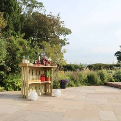 Elegant outdoor patio setup with a wooden bar cart, surrounded by lush greenery and blooming plants in a beautiful garden, perfect for outdoor entertaining and relaxing.
