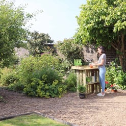 Lush garden with woman tending to plants on a wooden outdoor gardening station, surrounded by green trees, flowering plants, and natural landscape for outdoor living and gardening.