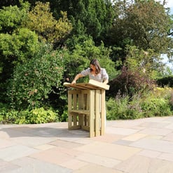 Impressive garden with lush greenery, flowering plants, and a woman enjoying outdoor reading at a custom wooden table, showcasing outdoor furniture and landscaping ideas from Empire Home and Garden.