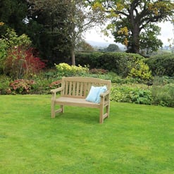 Peaceful garden with lush green grass, colorful shrubbery, and a wooden bench with cushions under tall trees, creating a perfect outdoor relaxing space.
