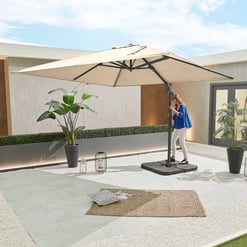 Stylish beige outdoor patio umbrella with a woman adjusting the height, surrounded by potted plants, decorative lanterns, and a cozy textured rug on a modern clean backyard space.