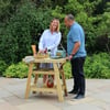 Fresh outdoor grilling and barbecue setup in a lush garden, featuring a wooden table with condiments, fresh ingredients, and a cheerful couple enjoying their time in nature.