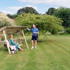 Natural outdoor garden swing set with a shaded canopy, surrounded by lush greenery and blooming flowers, perfect for relaxing and outdoor family enjoyment.