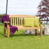 Natural wooden outdoor garden bench with colorful throw pillows and a purple blanket, situated on a well-maintained lawn with decorative rocks and lush greenery. Perfect for relaxing outdoor living spaces.