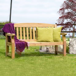 Natural wooden outdoor garden bench with colorful throw pillows and a purple blanket, situated on a well-maintained lawn with decorative rocks and lush greenery. Perfect for relaxing outdoor living spaces.