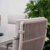Modern outdoor dining chair with beige woven backrest and armrest, set on a sleek white table with black plates, surrounded by lush green foliage and hydrangea plants.
