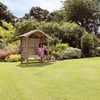 Large backyard garden with a woman sitting on a wooden garden bench under a small wooden gazebo, surrounded by lush green plants and flowering shrubs, ideal for outdoor relaxation.