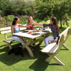 Relaxing outdoor garden dining scene with three women raising glasses of wine, surrounded by lush greenery and colorful flowers, on a sunny day at a wooden picnic table.