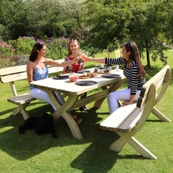 Relaxing outdoor garden dining scene with three women raising glasses of wine, surrounded by lush greenery and colorful flowers, on a sunny day at a wooden picnic table.