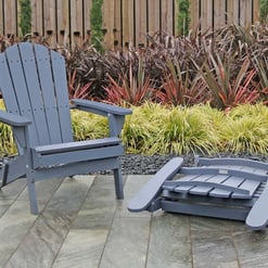 Blue Adirondack chair with matching fallen ottoman on a stone patio, surrounded by lush ornamental grasses and tall plants in a landscaped garden.