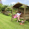 Relaxing woman sitting on a wooden garden bench with a canopy, surrounded by colorful flowers and lush greenery, enjoying outdoor leisure in a beautifully landscaped yard.