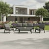 Contemporary outdoor patio furniture set with modern black cushions and matching table in front of a stylish modern home with lush landscaping and flowering bushes.