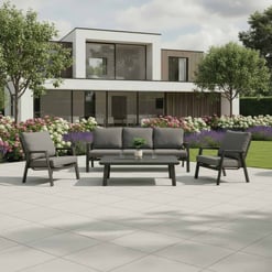 Contemporary outdoor patio furniture set with modern black cushions and matching table in front of a stylish modern home with lush landscaping and flowering bushes.