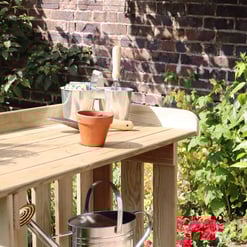 Rustic garden work table made of light wood with gardening tools, terracotta pot, and potted plants in a lush outdoor garden setting with blooming flowers and green foliage.