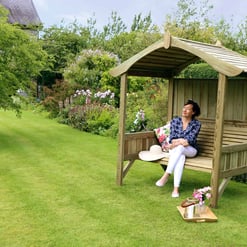 Relaxing garden sitting area with a woman enjoying the outdoor space, surrounded by vibrant flowers, lush greenery, and a wooden garden shelter for comfortable garden leisure and outdoor living.