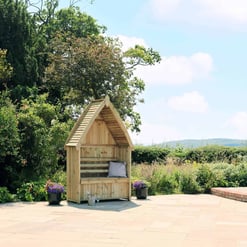 Wooden garden playhouse with bench and pillows on a stone patio surrounded by lush greenery and colorful flowers, ideal for outdoor relaxation and garden decor.