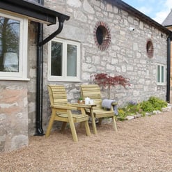 Cozy outdoor seating area with wooden chairs and a table next to a stone house, surrounded by garden plants, enhancing home and garden aesthetic.