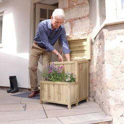 Raised wooden planter box outdoor garden decor with blooming flowers in front of a rustic stone and siding exterior wall. Perfect for enhancing curb appeal and outdoor gardening.