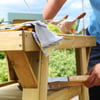 An outdoor wooden table being assembled, suitable for gardening or outdoor dining, featuring a natural wood finish with tools and ingredients for a fresh salad on a sunny day.