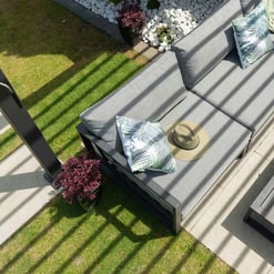 Modern outdoor patio seating area with gray cushioned sectional, decorative pillows, sun hat, surrounded by lush landscaping and white river rocks, perfect for garden and backyard entertainment.